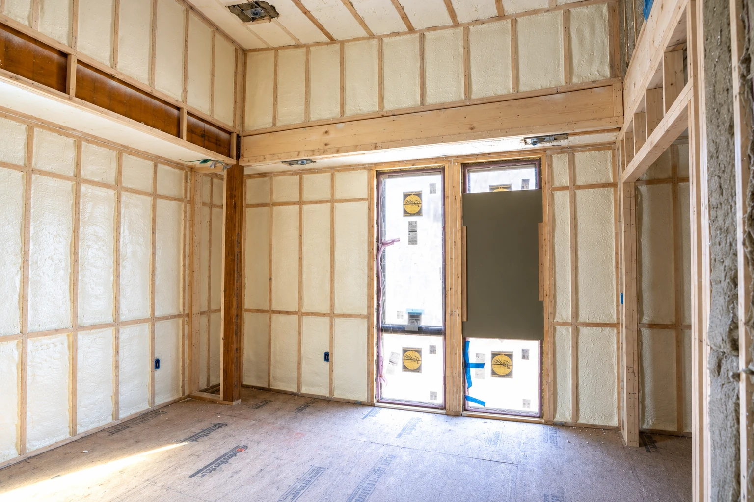 An unfinished room has pale spray foam insulation between exposed wooden studs and across the ceiling.
