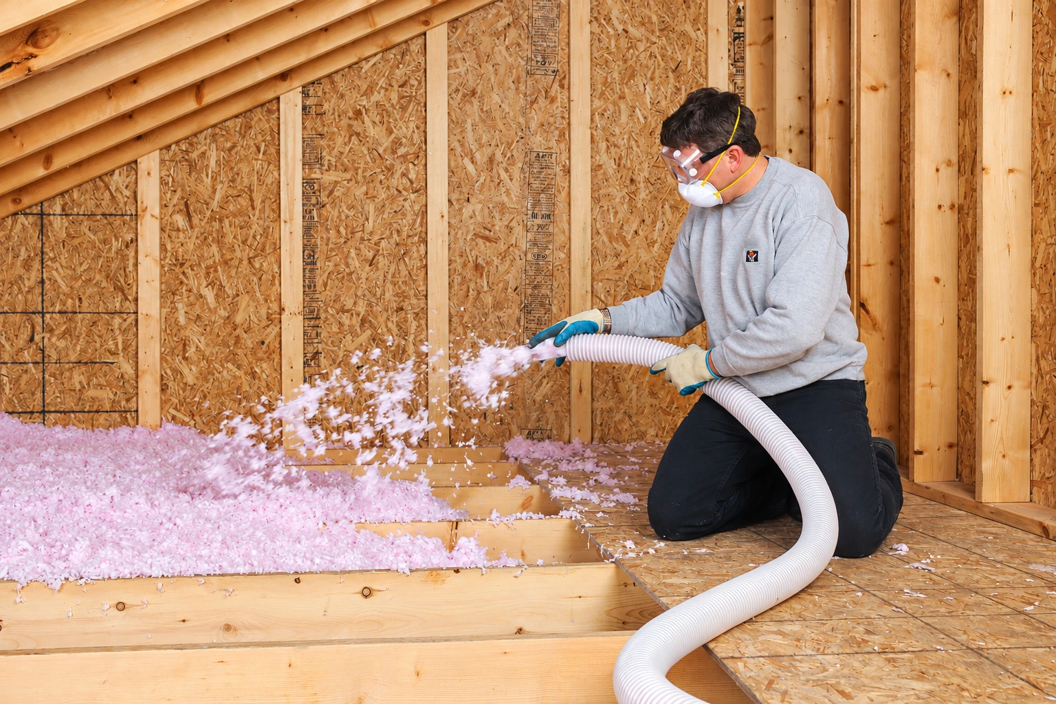 A kneeling worker sprays pink blown-in insulation through a hose across an unfinished attic floor.