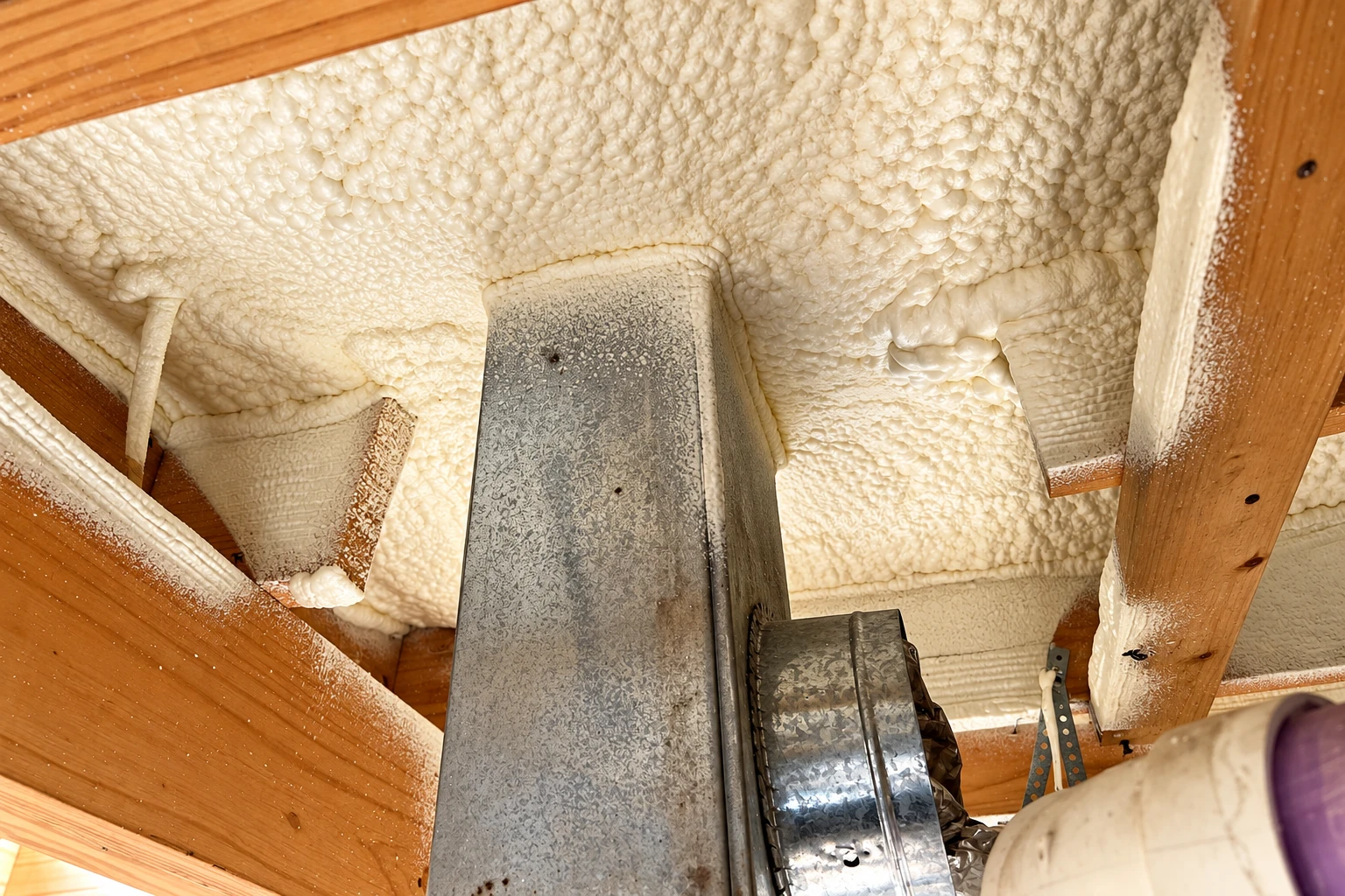 Spray foam insulation covers the underside of a crawl space floor around a large metal duct and exposed wood joists.