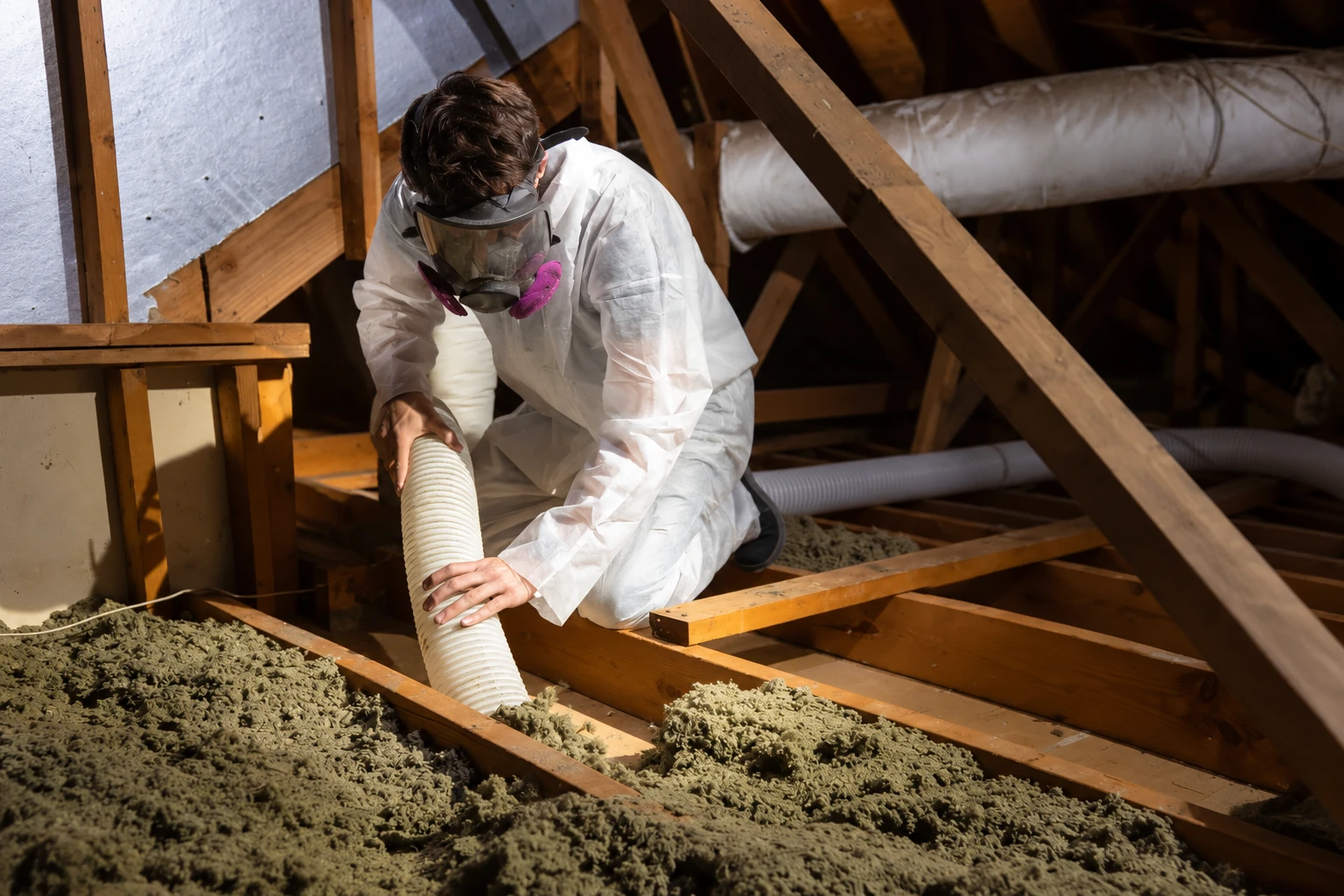 A worker in protective coveralls vacuums loose attic insulation through a hose among exposed wooden rafters.