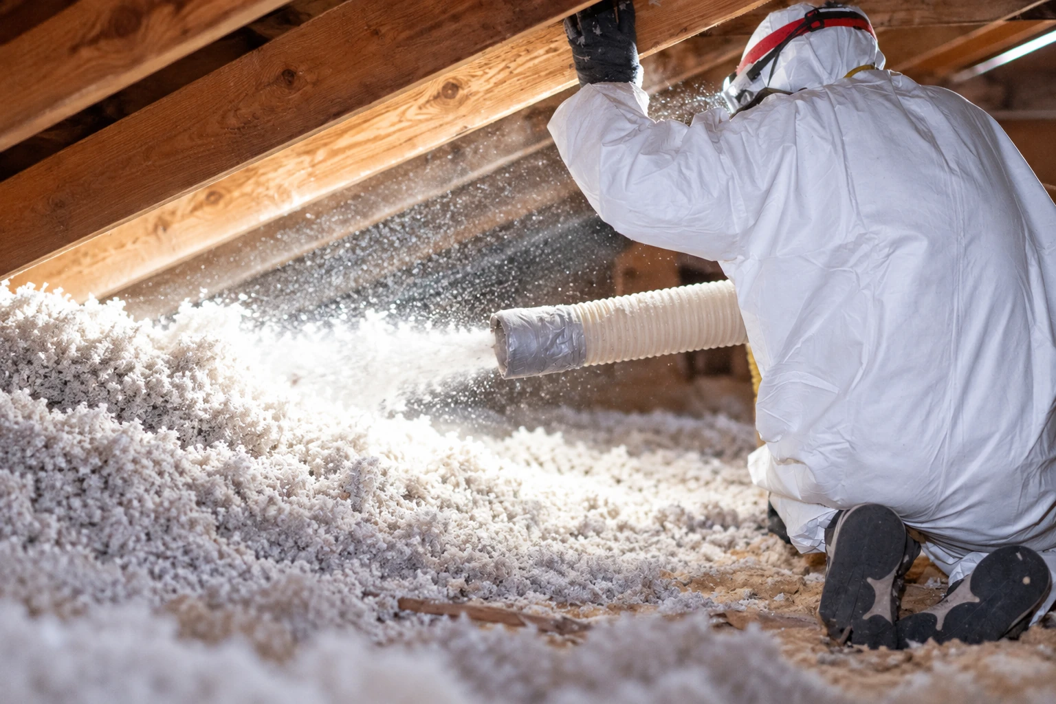 A worker in protective coveralls sprays loose white insulation through a hose across an attic floor beneath exposed rafters.